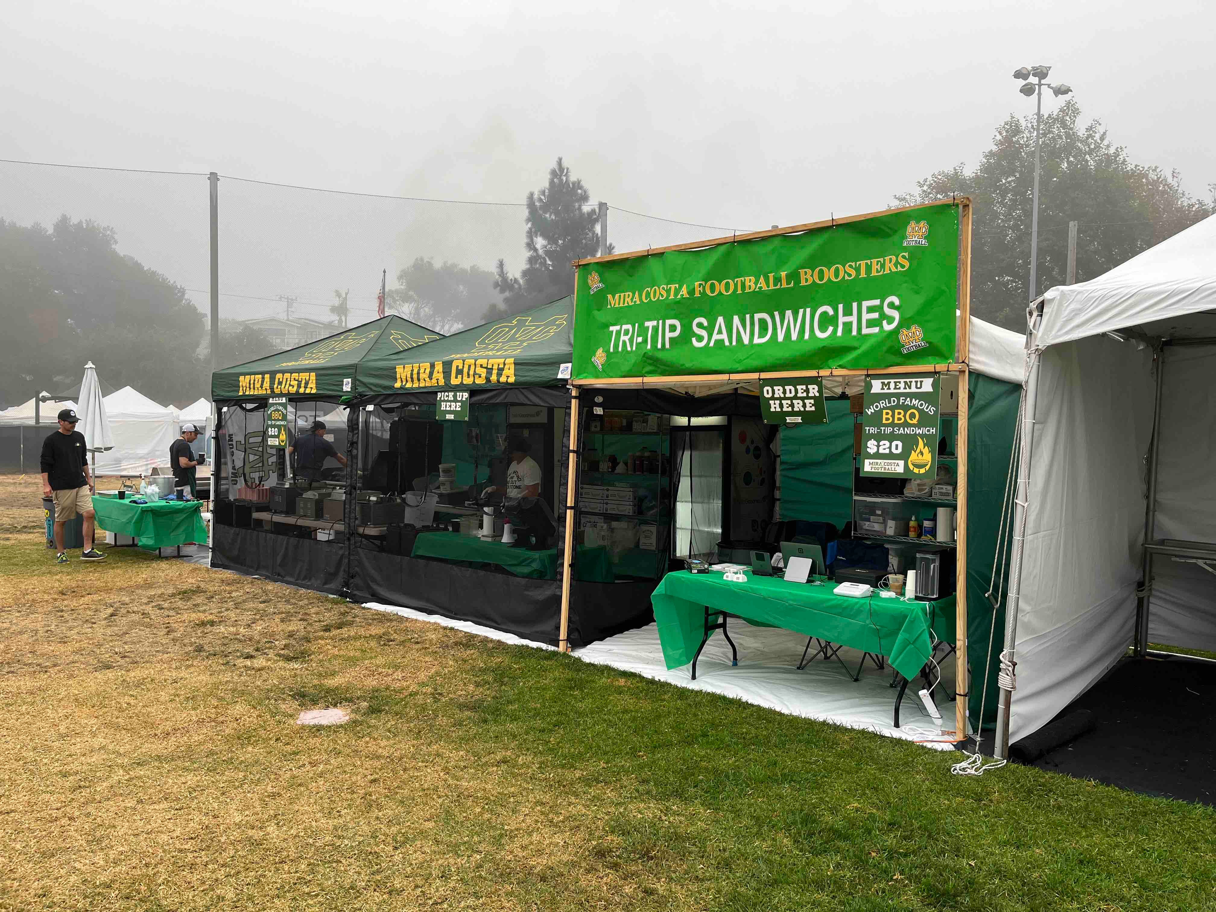 The Mira Costa Tri-Tip Booth at the Manhattan Beach Hometown Fair — green canopies, the $20 tri-tip sandwich menu, and the crew at work